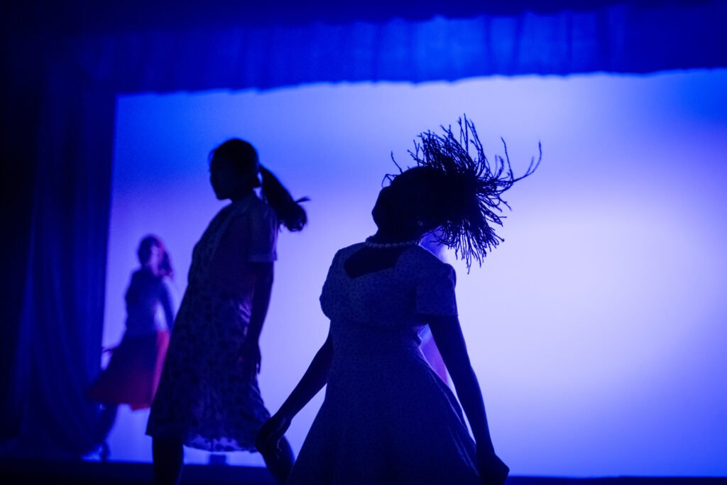 Silhouettes of young female vocalists or dancers performing dynamically under vibrant blue stage lighting during a student live music showcase.
