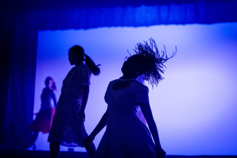 Silhouettes of young female vocalists or dancers performing dynamically under vibrant blue stage lighting during a student live music showcase.