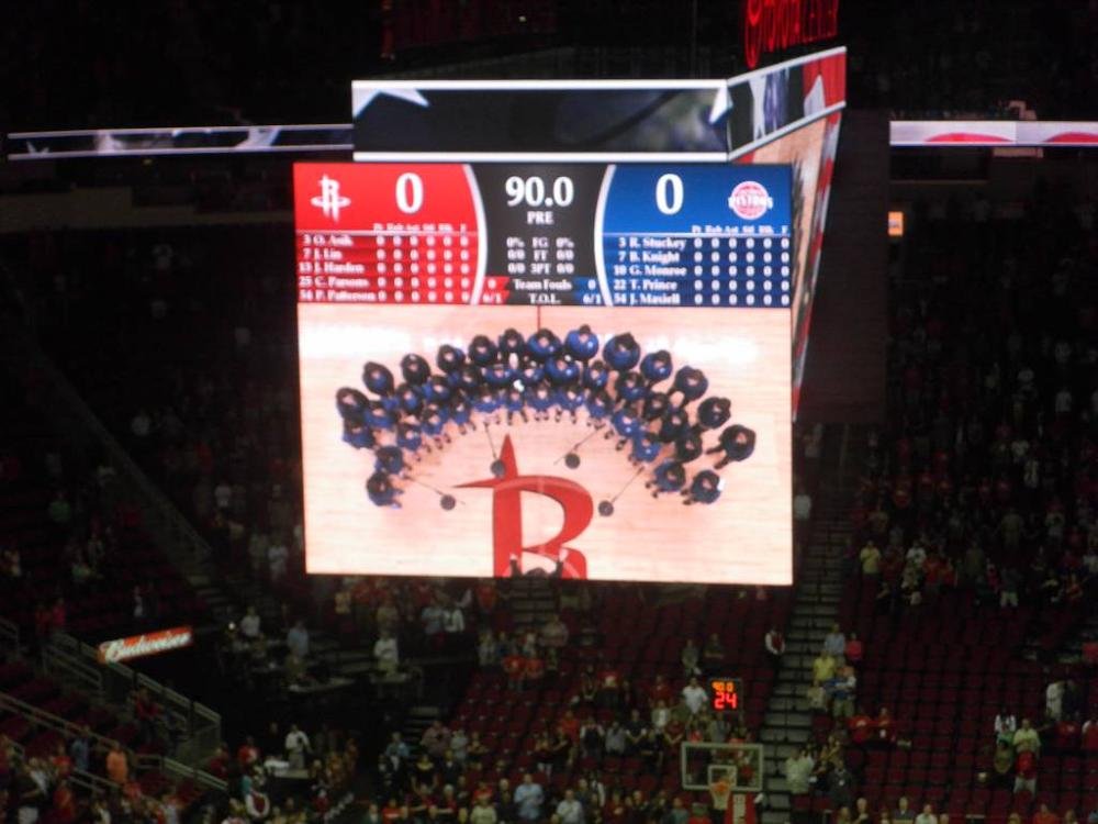 A massive stadium jumbotron screen inside the Toyota Center showing a live pre-game dance team performance over the Houston Rockets center-court logo.